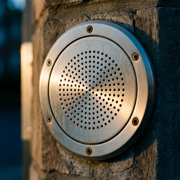 Brushed stainless intercom speaker grille mounted on a stone gate pillar in warm dusk light