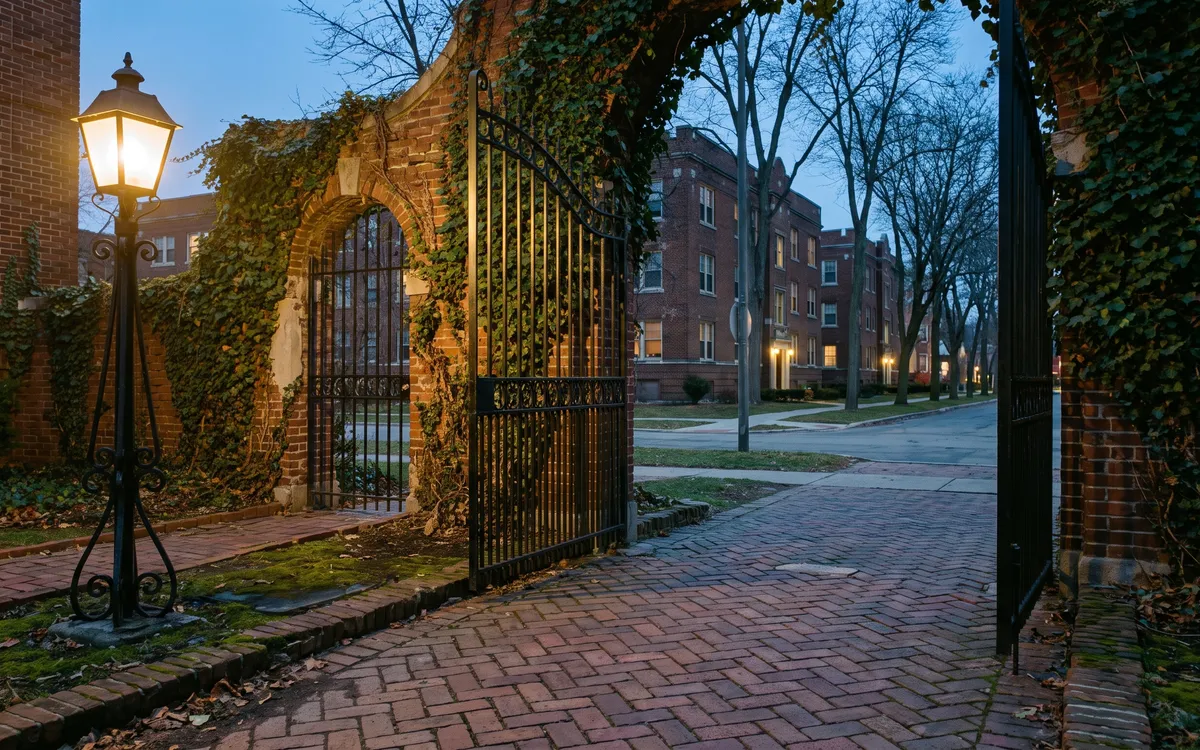 Black iron gate in an ivy covered brick archway beside a glowing post lantern on a blue hour Ann Arbor street of brick apartment houses