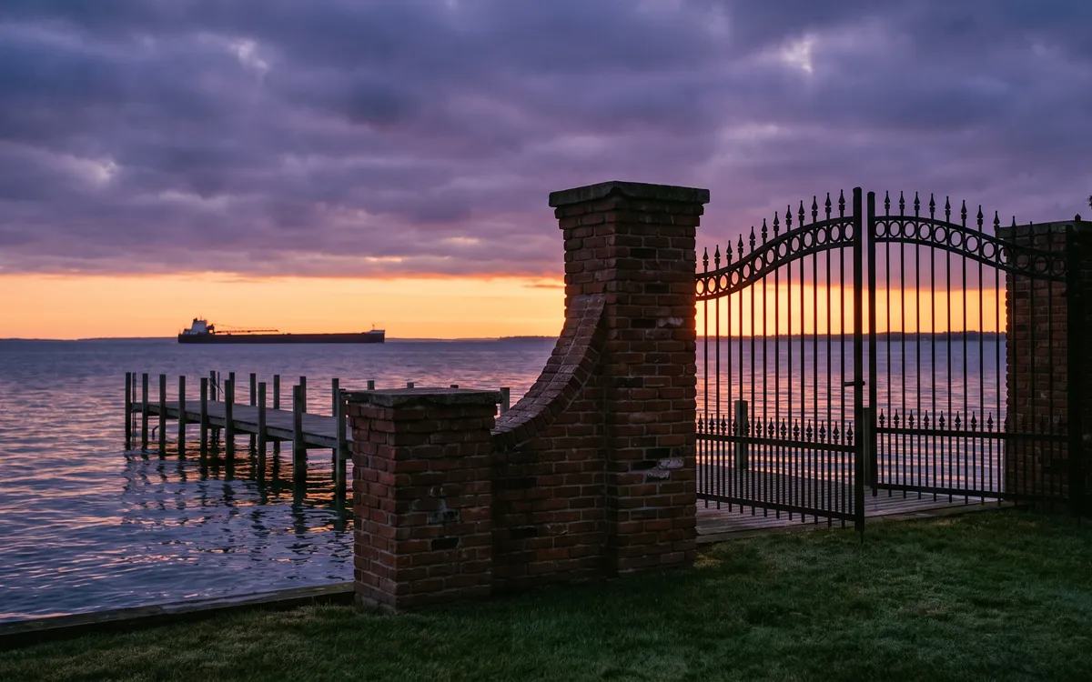 Iron estate gate and brick pillars at the water edge with a private pier and a distant freighter silhouette on the lake in Grosse Pointe Park at deep dusk